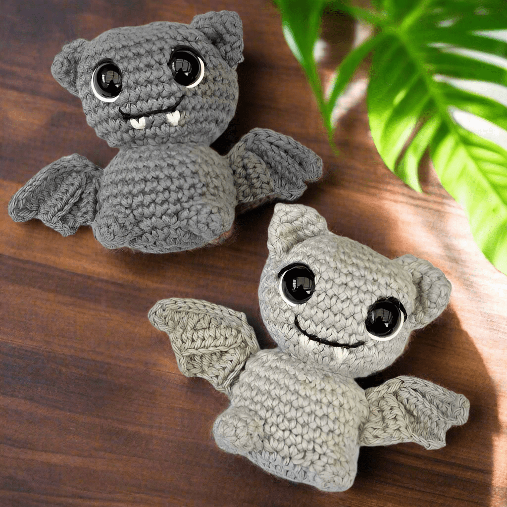 Two crocheted gray bat plushies with large eyes on a wooden surface with green leaves