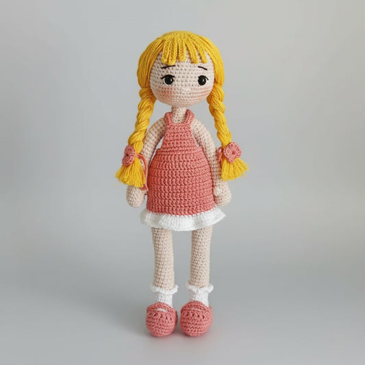 Crocheted doll with yellow braids, pink dress, and white ruffled socks on gray background