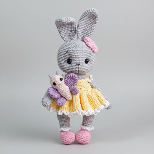 Crocheted gray bunny in yellow dress holding purple stuffed toy, wearing pink bow and shoes