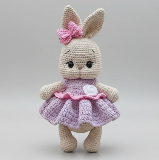Crocheted bunny with pink bow and lavender dress, standing against neutral background