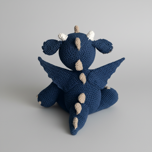 Blue crocheted dragon with tan spikes, sitting on a light gray background.