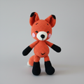 Crocheted orange fox stuffed animal with black ears and paws, standing upright.