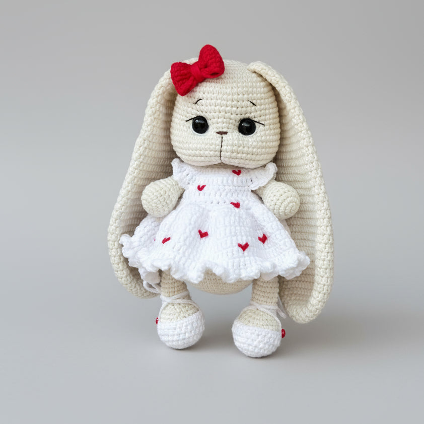 Crocheted white bunny with red bow and heart-dotted dress on gray background