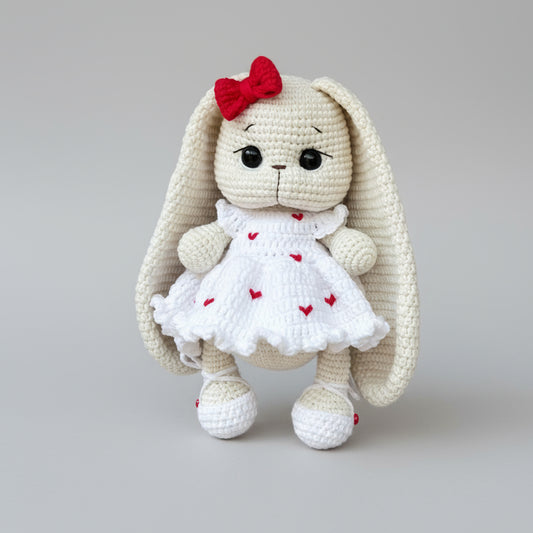 Crocheted white bunny with red bow and heart-dotted dress on gray background