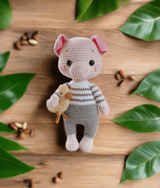 Crocheted pig plushie in striped sweater, holding a small yellow bird, surrounded by green leaves