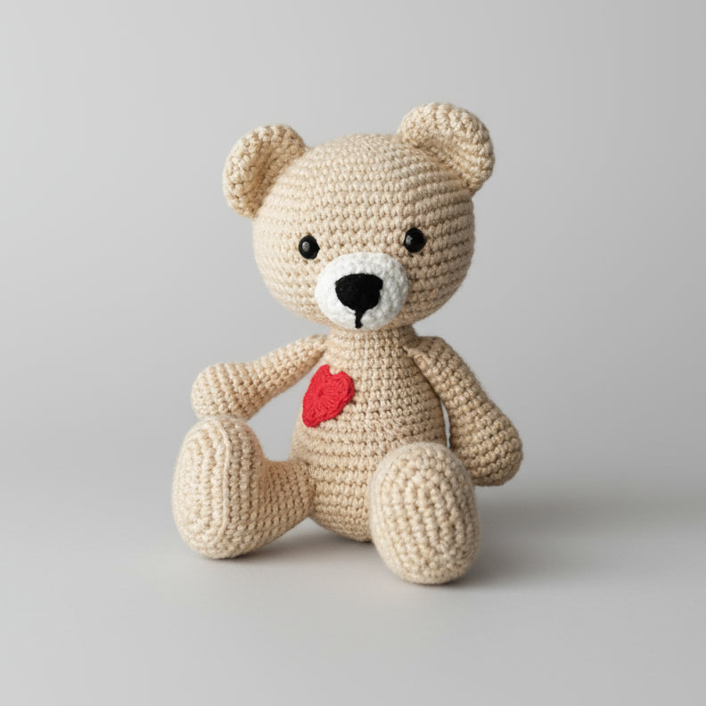Crocheted beige teddy bear with red heart, sitting against neutral background
