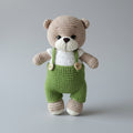 Crocheted teddy bear in green overalls standing against a gray background