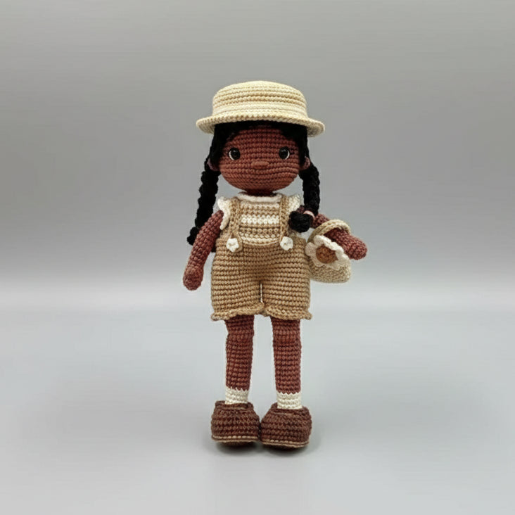 Crocheted doll with braids, straw hat, overalls, and basket in neutral tones