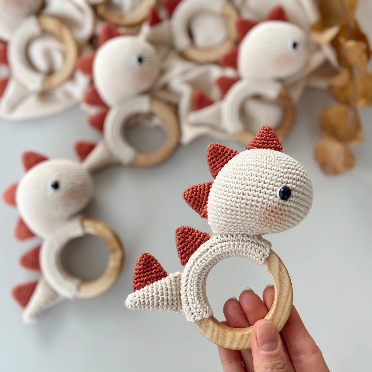 Crocheted white and red dinosaur teething ring held by a hand, with similar toys in background