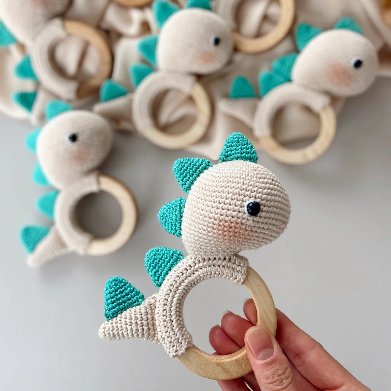 Crocheted teal and white dinosaur-shaped baby teething ring held by a hand