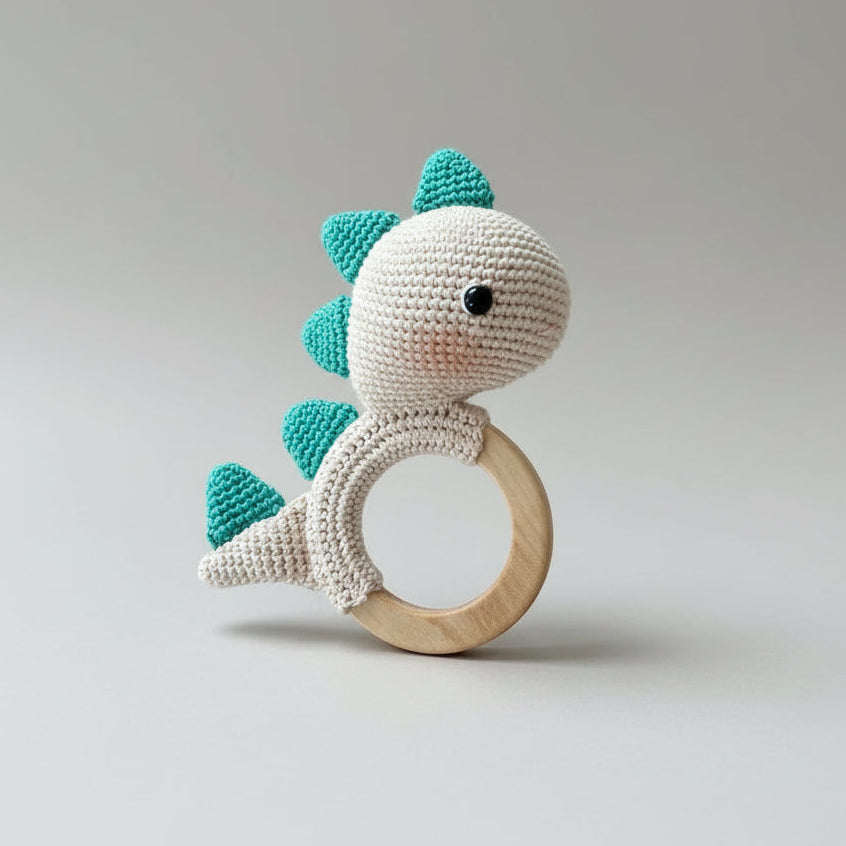 Crocheted teal and white dinosaur-shaped baby teething ring held by a hand