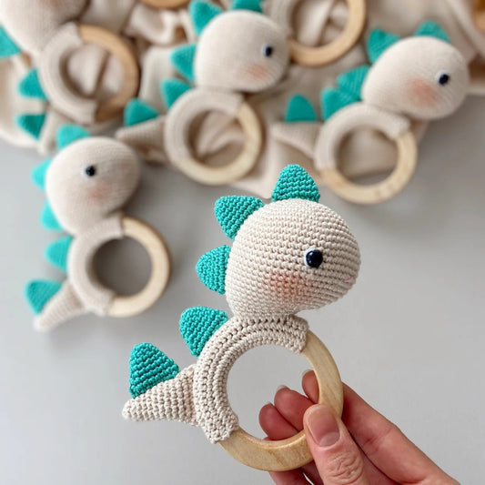 Crocheted teal and white dinosaur-shaped baby teething ring held by a hand