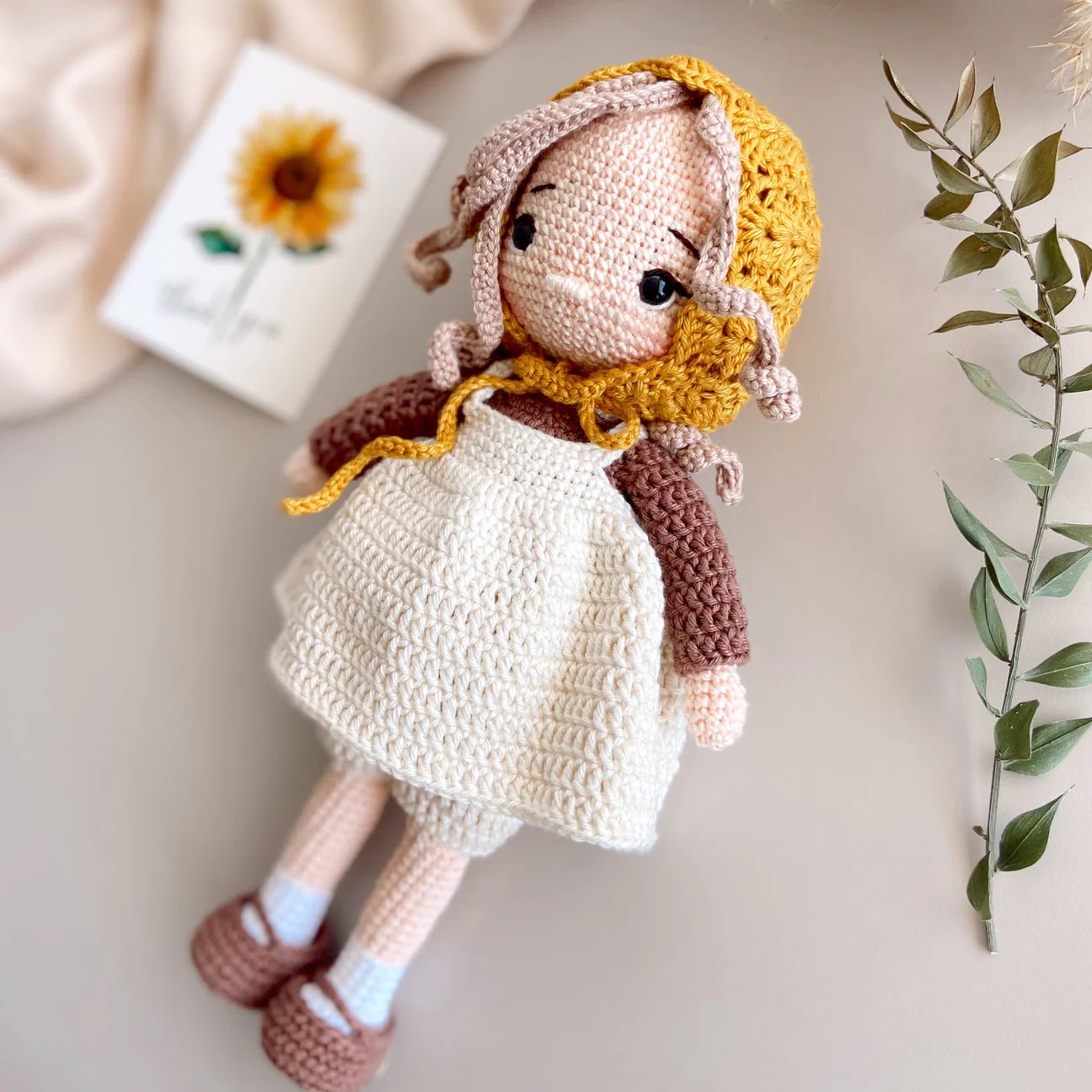 Crocheted doll with yellow bonnet, white dress, and brown shoes, sitting near a sunflower.
