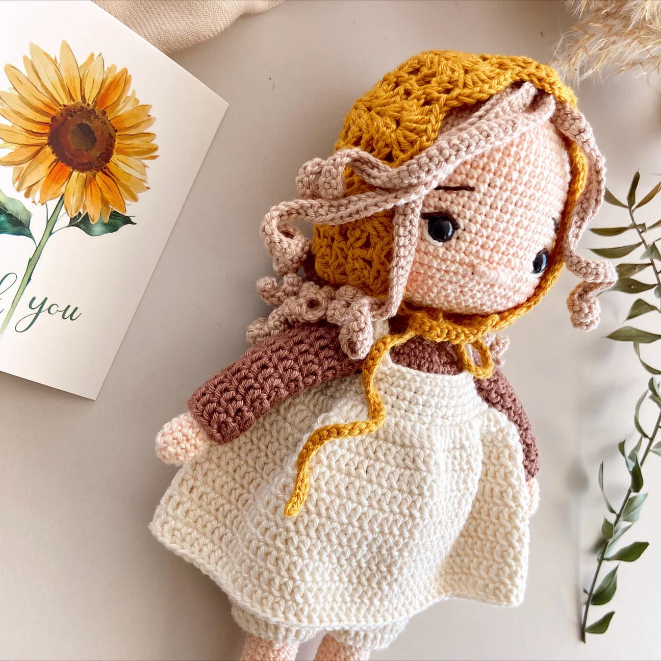 Crocheted doll in white dress with yellow bonnet, beside a sunflower greeting card
