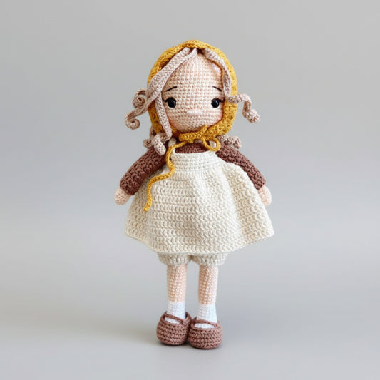 Crocheted doll with yellow bonnet, white dress, and brown shoes standing against gray background