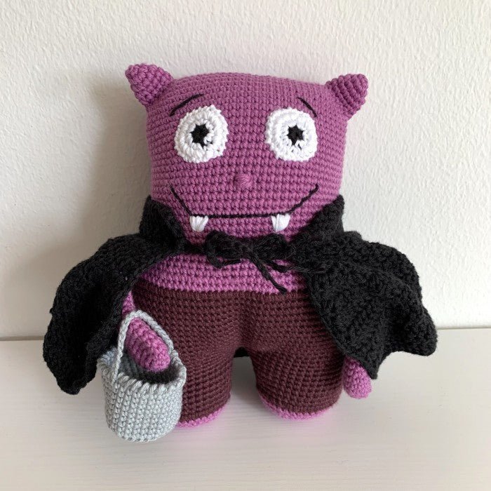 Purple crocheted monster with black wings, holding a gray bucket, smiling with fangs.