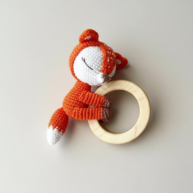 Crocheted orange fox holding a wooden teething ring against a neutral background