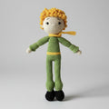 Crocheted doll with yellow curly hair, green outfit, yellow scarf, and black shoes, standing on a neutral background.