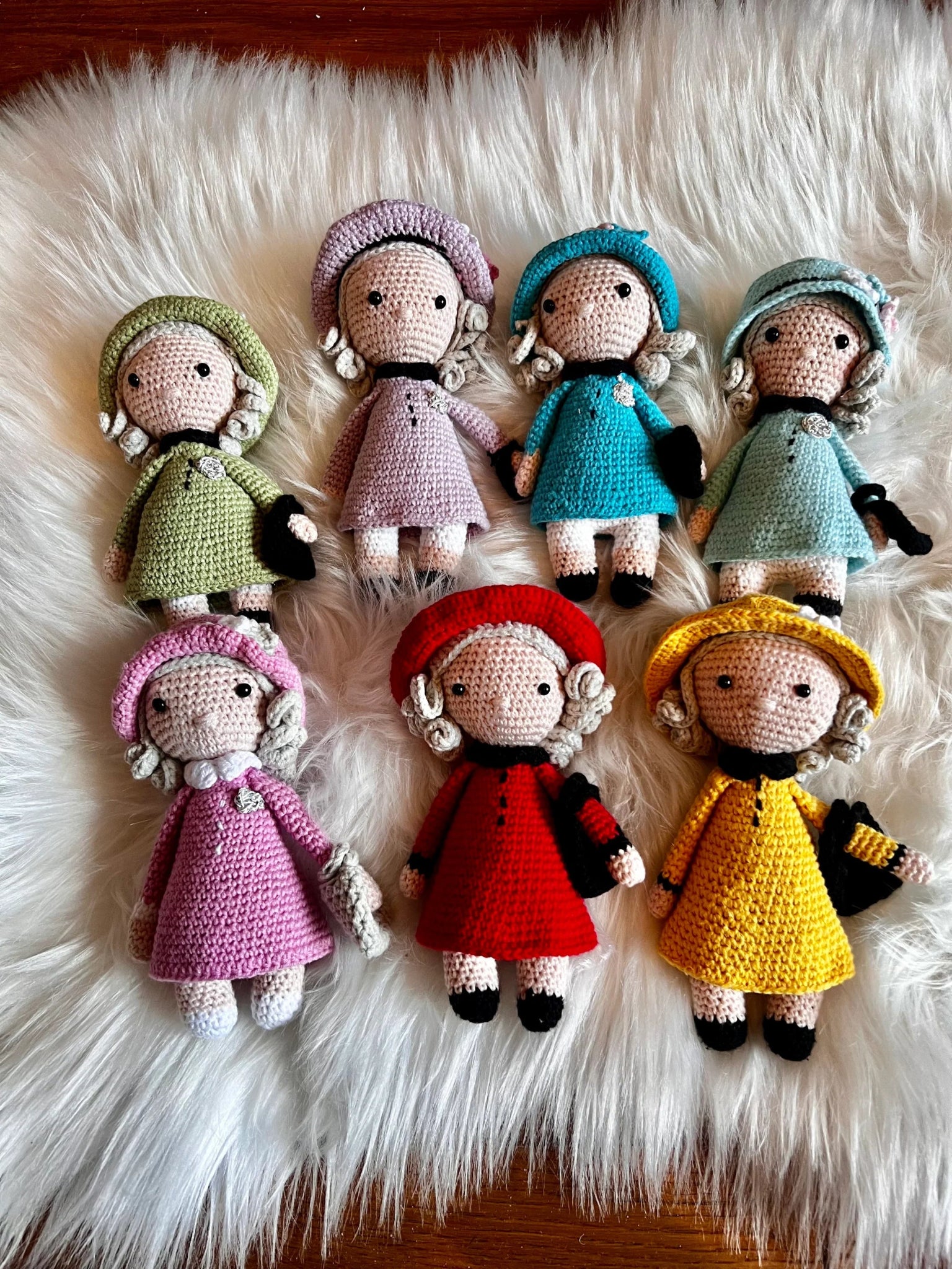 Six crocheted dolls in different colored dresses and bonnets, arranged on a fluffy white fur background with vintage-inspired styling.