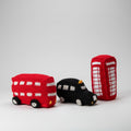 Crocheted red double-decker bus, black taxi, and red phone booth sit on windowsill with garden and trees visible through window.