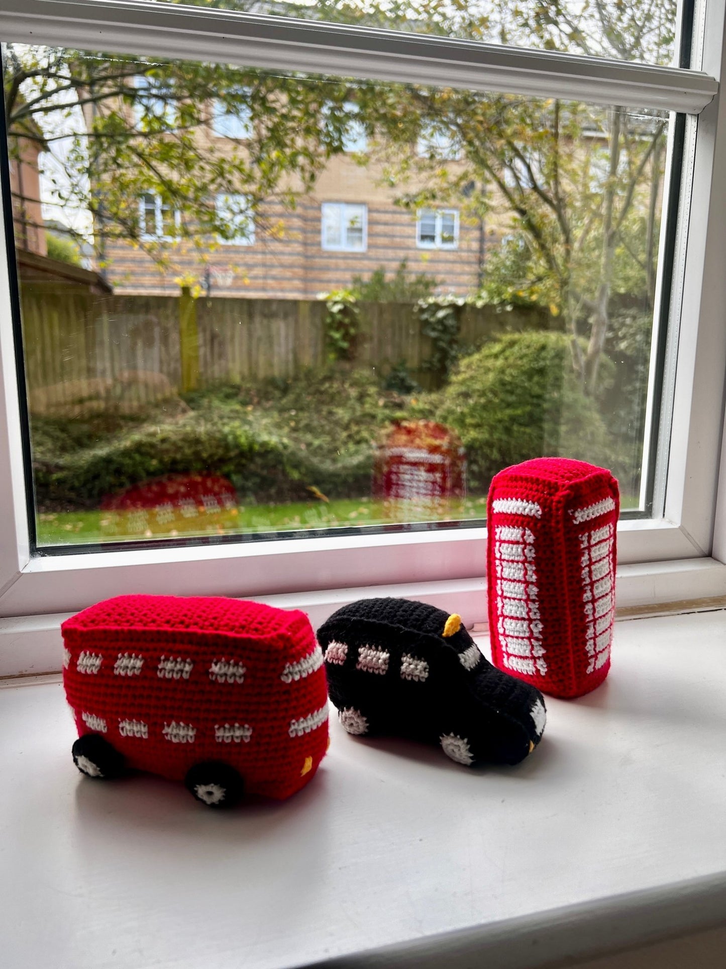 Crocheted red double-decker bus, black taxi, and red phone booth sit on windowsill with garden and trees visible through window.