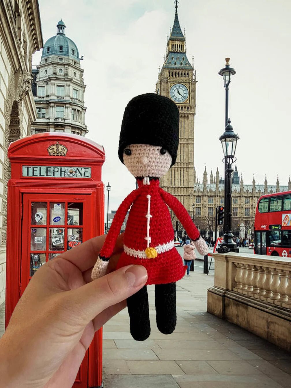 Crocheted royal guard figurine held in front of London landmarks, including red telephone booth, Big Ben, and red double-decker bus.