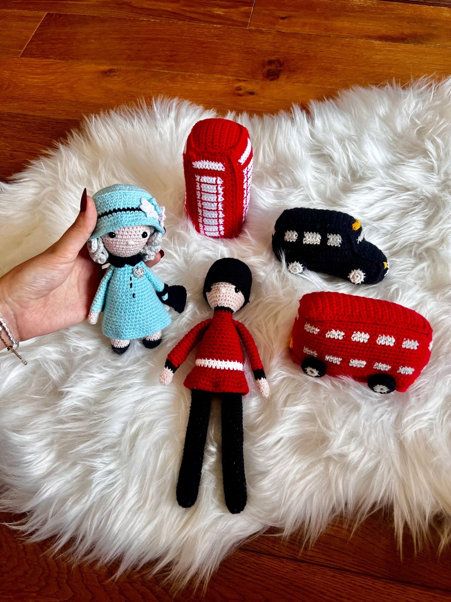 Handmade crochet dolls and London-themed objects on white fur, including a blue-dressed doll, red phone booth, black taxi, and red bus.