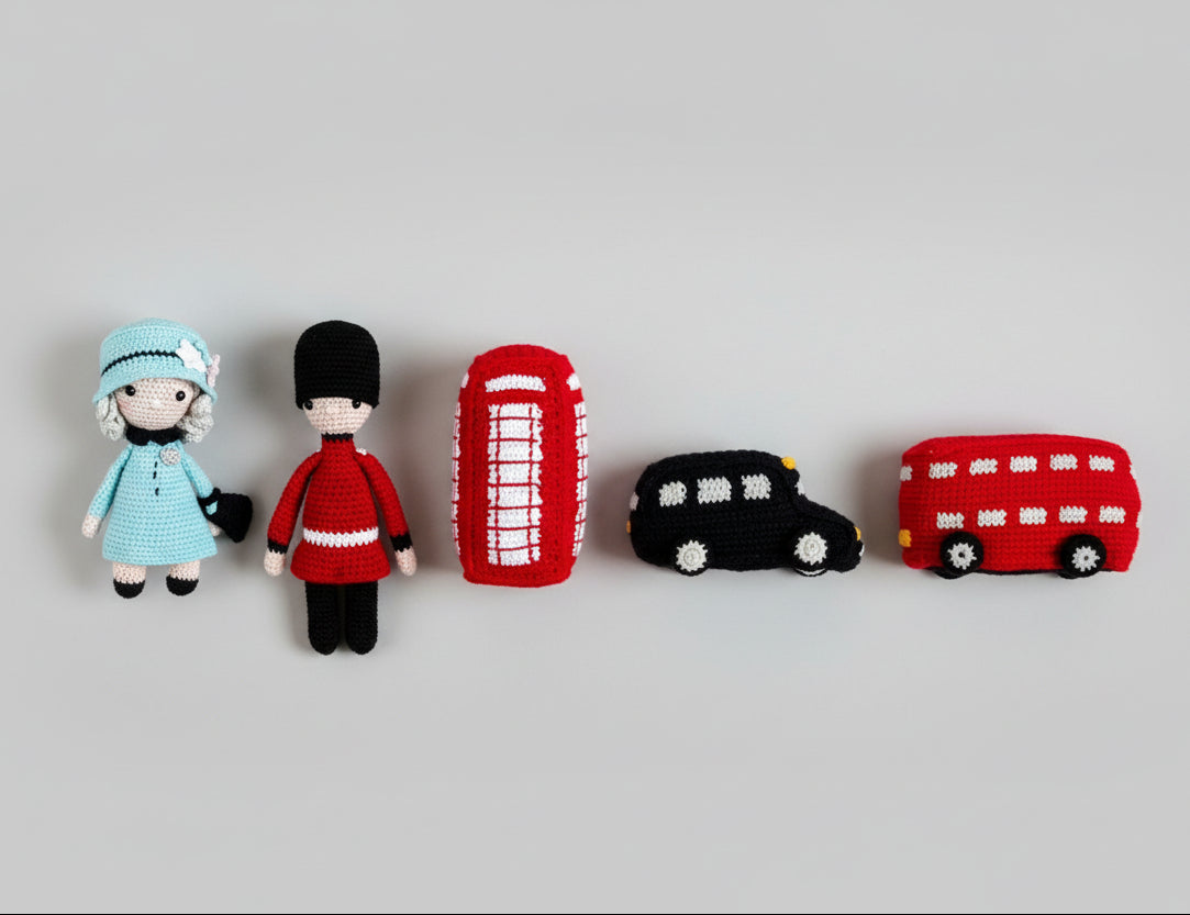 Handmade crochet dolls and London-themed objects on white fur, including a blue-dressed doll, red phone booth, black taxi, and red bus.