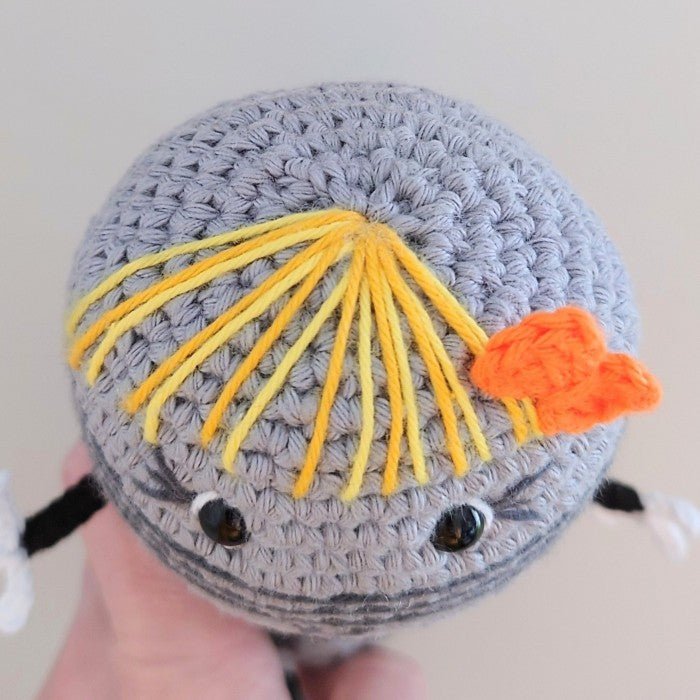 Crocheted gray whale with yellow yarn hair and orange tail fin, held in hand