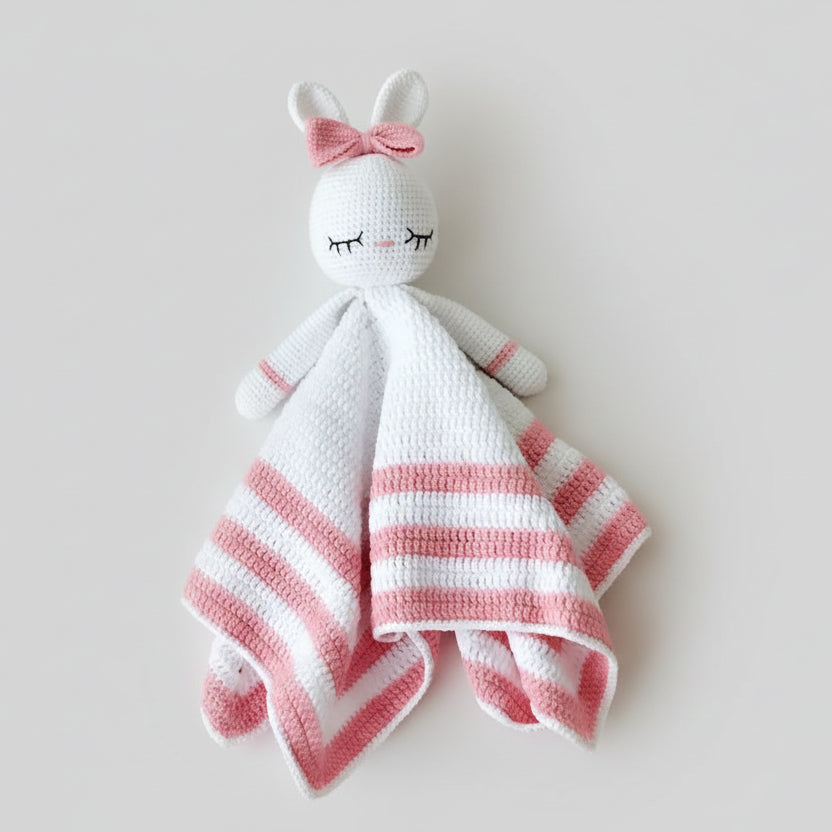 White and pink crocheted bunny lovey with closed eyes and a bow on soft fabric