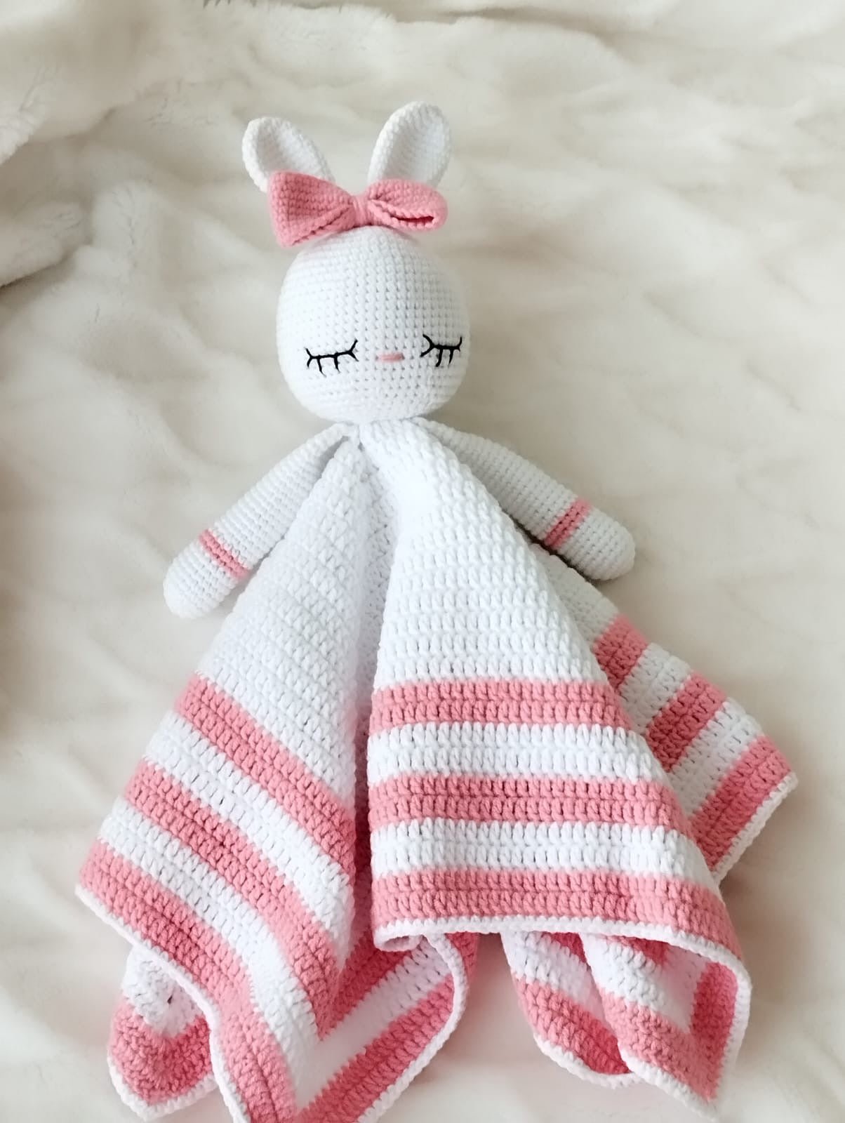 White and pink crocheted bunny lovey with closed eyes and a bow on soft fabric
