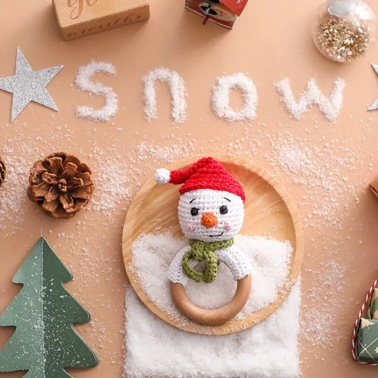Crocheted snowman baby rattle on wooden plate with scattered snow and holiday decorations