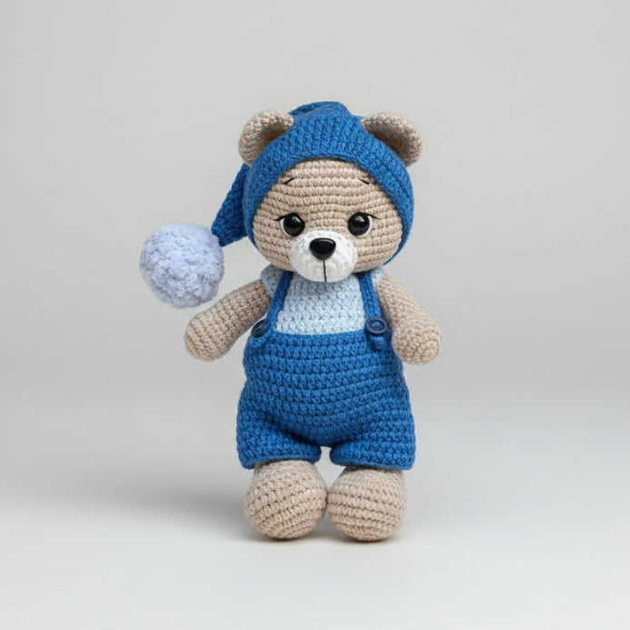 Crocheted teddy bear in blue overalls and nightcap, standing against gray background
