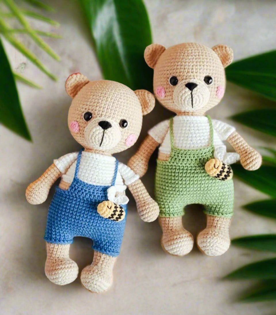 Two crocheted teddy bears wearing blue and green overalls, surrounded by green leaves.