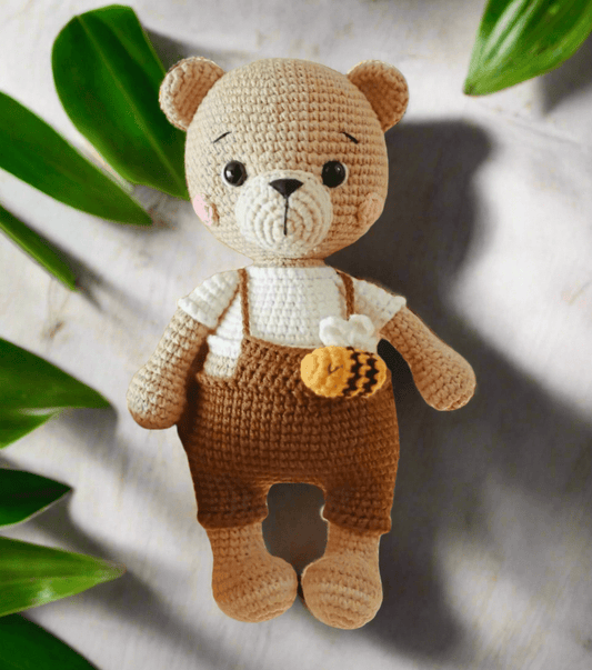Crocheted teddy bear with brown overalls and honey bee, surrounded by green leaves