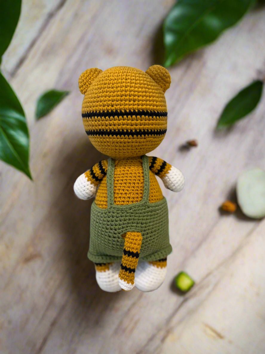 Crocheted tiger-striped teddy bear in green overalls, sitting on wooden surface