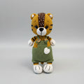 Crocheted tiger plush wearing green overalls, standing against gray background