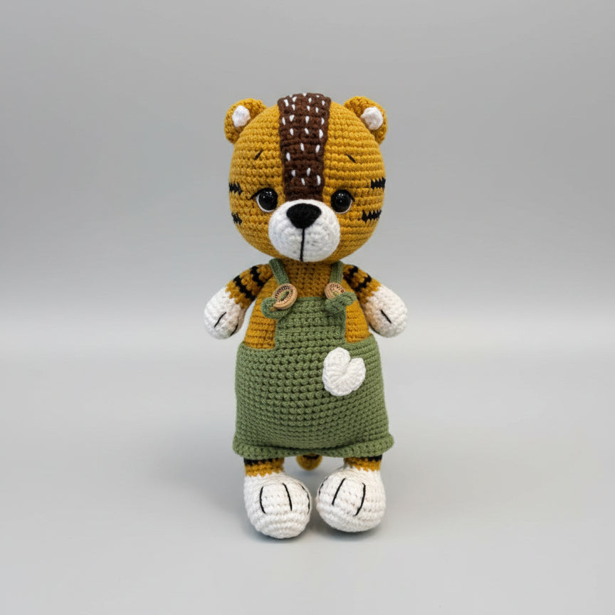 Crocheted tiger plush wearing green overalls, standing against gray background