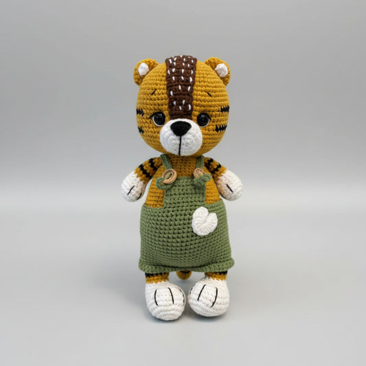 Crocheted tiger plush wearing green overalls, standing against gray background