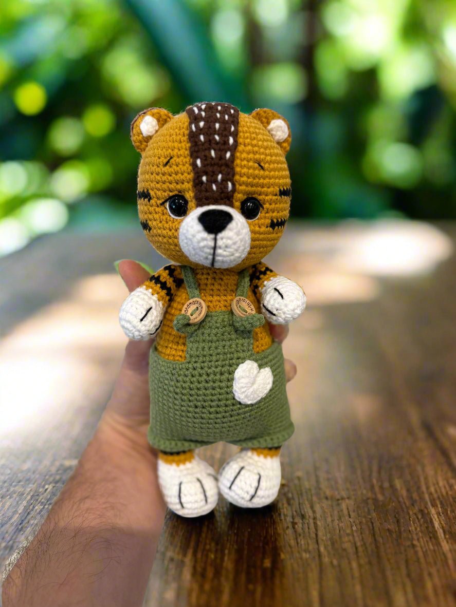 Crocheted tiger stuffed animal in green overalls, held in hand against blurry background