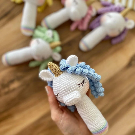 Crocheted white and blue unicorn rattle with golden horn, held in hand over wooden surface