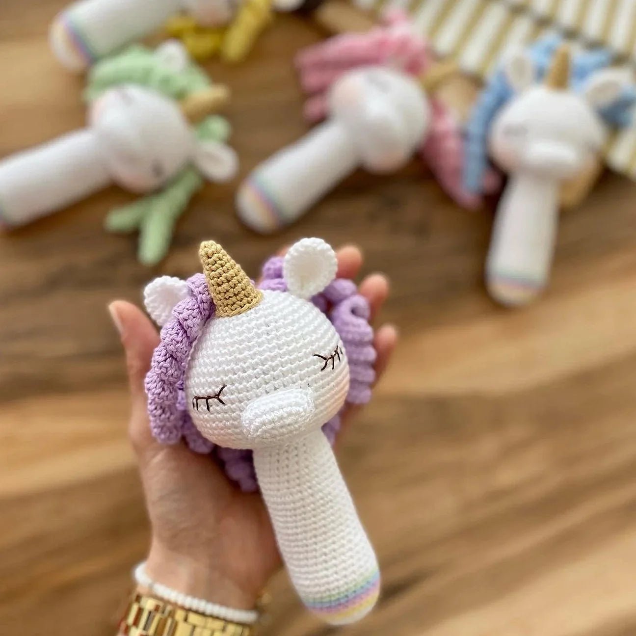 Crocheted unicorn rattle in white, purple, and gold, held in a hand with blurred similar toys