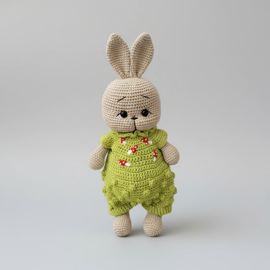 Crocheted bunny in green overalls with red mushroom embroidery, standing against gray background