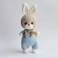 Crocheted bunny in blue overalls, holding a tiny carrot, against a soft gray background