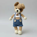 Crocheted teddy bear in blue overalls with paw print, standing on gray background