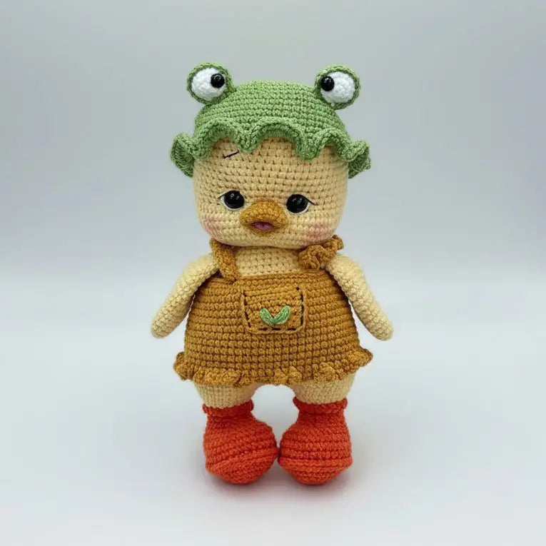 Crocheted chick doll with frog hat, yellow and orange, standing on neutral background