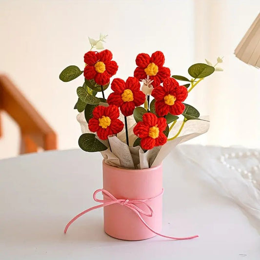 Crocheted red flowers with yellow centers in a pink tied container with green leaves