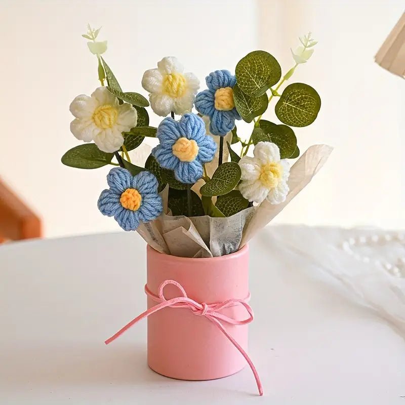 Crocheted white and blue flowers in a pink vase with a tied bow, sitting on a white surface.