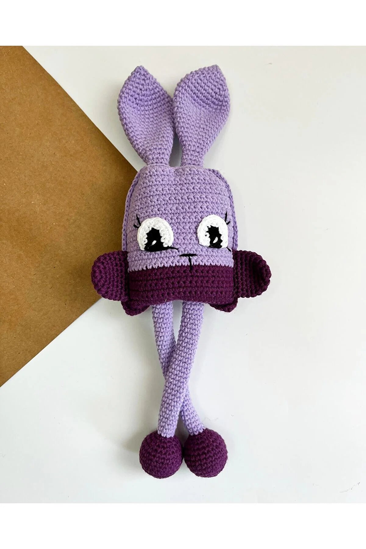 Crocheted purple bunny with long ears and large eyes, sitting against white and brown background