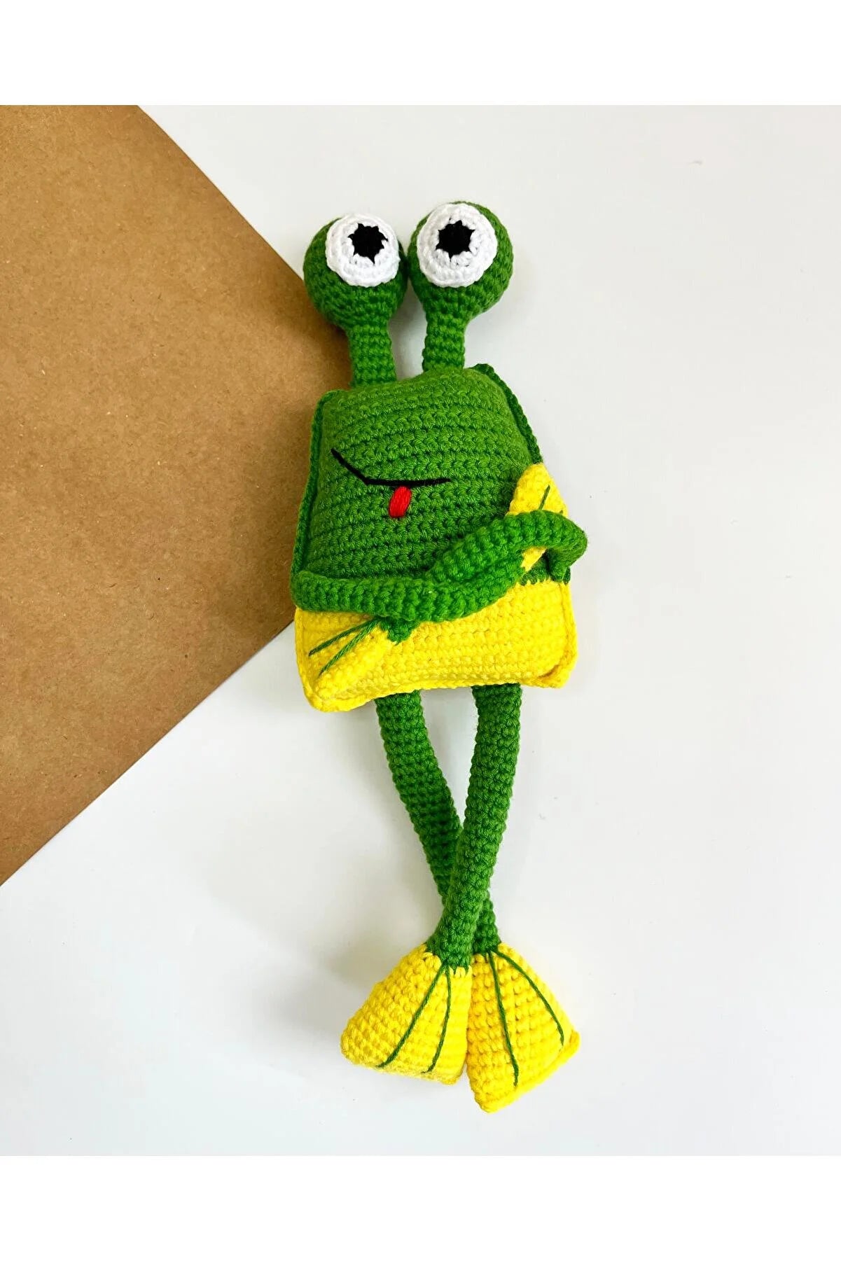 Green crocheted frog toy with bulging eyes and yellow feet, sitting playfully